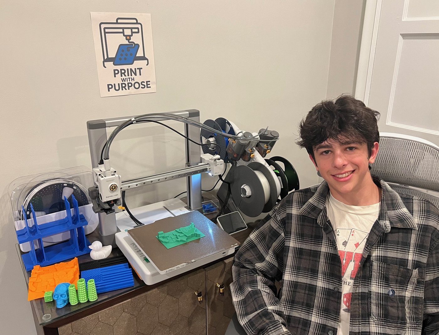 Jackson Teitelbaum next to his 3D printer and a table of tactile prints, under a Print with Purpose sign